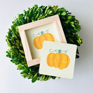 May include: A small wooden frame with a white background and a hand-painted orange pumpkin with a green stem. The frame is sitting on top of a green boxwood wreath.
