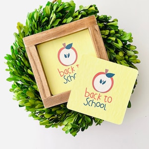 May include: A yellow wooden sign with a red apple graphic and the text "back to school" in blue and red lettering. The sign is sitting on a green wreath.