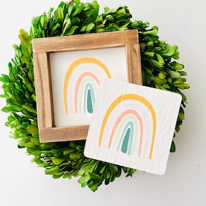 Puede incluir: Dos piezas de decoración de pared con temática de arcoíris. Una es un letrero de madera enmarcado con un diseño de arcoíris en tonos de amarillo, naranja, rosa, azul y verde. La otra es un letrero más pequeño, sin marco, con un diseño de arcoíris similar.