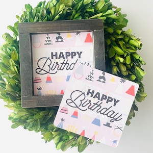 Könnte beinhalten: Zwei kleine Schilder mit einem rosa und weißen Hintergrund mit bunten geometrischen Formen und dem Text "Happy Birthday" in Schwarz. Die Schilder sind in Holz gerahmt.