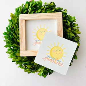 May include: A square wooden framed sign and a coaster featuring a smiling yellow sun with the text "you are my sunshine" in cursive. The sign and coaster are set on a green leafy wreath.