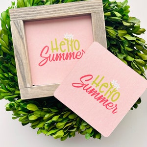 May include: Two pink square coasters with the text "Hello Summer" in green and red lettering. The coasters are on top of a green bush.