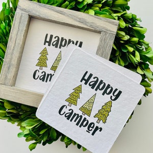 May include: Two white square signs with black text that reads "Happy Camper" and three green stylized pine trees. The signs are on a white background.