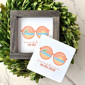 May include: A square wooden framed print and a square coaster feature a design of orange sunglasses reflecting a beach scene with the text "sunshine on my mind". The items are surrounded by a green wreath.