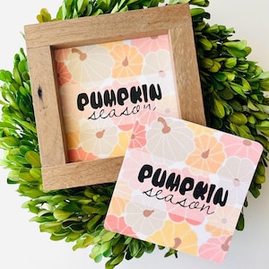 May include: Two framed signs with a pumpkin pattern and the text "Pumpkin season" in black lettering. The signs are on a green wreath.