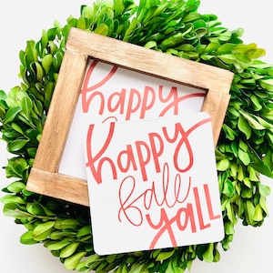 May include: A white wooden sign with the words "Happy Fall Y'all" written in red cursive lettering. The sign is sitting on a green boxwood wreath.