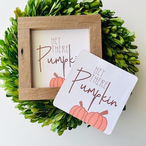 May include: Two wooden signs with the text "Hey there Pumpkin" in brown lettering and a pink pumpkin illustration. The signs are on a green leafy background.