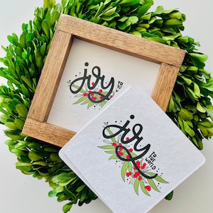 May include: Two white square signs with black calligraphy that says "joy" with a green leafy branch and red berries. The top sign says "joy to" and the bottom sign says "joy to the world".