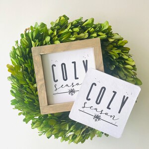 May include: Two white signs with black text that reads "Cozy Season" with a snowflake graphic. The signs are sitting on a green boxwood wreath.