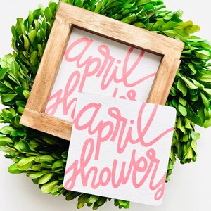Puede incluir: Un letrero de madera blanco con letras rosas que dice "April Showers". El letrero está sobre una corona verde.