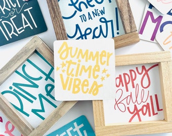 May include: A collection of wooden signs with various hand-lettered phrases, including "Cheers to a New Year!" , "Summer Time Vibes", "Happy Fall Y'all", "Sweet Land of Liberty", and "Merry and Bright".