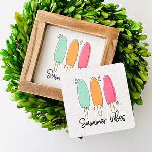 May include: A wooden framed sign and a coaster featuring colorful popsicle illustrations in shades of green, orange, and pink. The text "Summer Vibes" is written in black script. The items are surrounded by a wreath of green leaves.