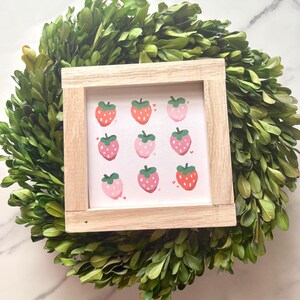 May include: A square wooden framed print featuring nine strawberry illustrations in shades of red and pink. The print is set against a background of a green leafy wreath.