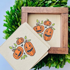 Puede incluir: Tres calabazas naranjas en forma de jack-o'-lantern con ojos y bocas negras están dibujadas sobre un fondo beige. Las calabazas están rodeadas de hojas verdes y corazones negros. La imagen está en un marco de madera.