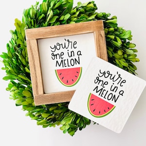 May include: Two wooden signs with the text "you're one in a melon" and a watermelon graphic. The signs are on a green leafy background.