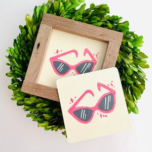May include: Two small framed illustrations of pink sunglasses with black lenses on a white background. The frames are decorated with small black dots.