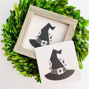 May include: A framed black and white illustration of a witch's hat with a black belt and stars. The frame is made of wood and has a light brown finish.