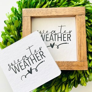 May include: Two white signs with black text and a heart design. The signs read "sweater weather". One sign is in a wooden frame.