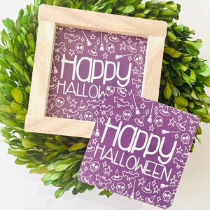 May include: Purple square sign with white text that reads "Happy Halloween" and a pattern of white skulls, stars, witches hats, and broomsticks. The sign is in a wooden frame.
