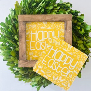 May include: A yellow wooden sign with white text that reads "Happy Easter" surrounded by a white pattern of eggs, chicks, and bunnies. The sign is sitting on top of a green boxwood wreath.