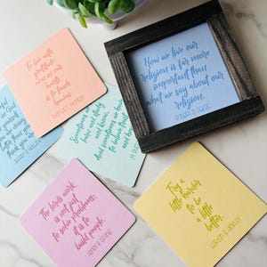 May include: A collection of square coasters with inspirational quotes in various colors, including peach, blue, and yellow. A framed quote is also present. The coasters feature quotes from religious figures.