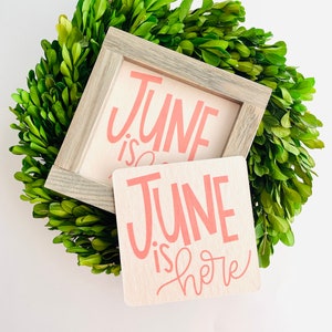 May include: Two wooden signs with the text "June is here" in pink lettering. The signs are sitting on top of a green boxwood wreath.