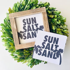 May include: Two white signs with black text and palm tree silhouettes. The signs read "SUN SALT SAND". The signs are on a green leafy background.
