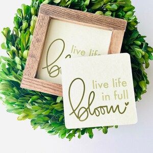 Peut inclure: Deux pancartes en bois avec le texte "live life in full bloom" écrit en lettres d'or. Les pancartes sont posées sur une couronne de buis verte.