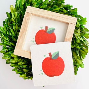 Op de afbeelding: Een ingelijst afdruk van een rode appel met een groen blad op een witte achtergrond. De lijst is gemaakt van licht hout en ligt op een groene krans.