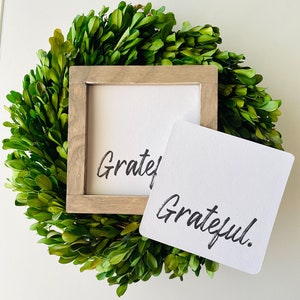 May include: A wooden framed sign with the word "Grateful" written in black calligraphy. The sign is sitting on top of a green boxwood wreath.