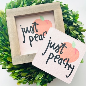 May include: A square wooden sign and a coaster with a peach graphic and the words "just peachy" in black script. The sign is in a grey frame, and both items have a light pink background. The items are surrounded by a green wreath.