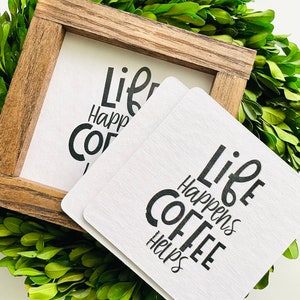 Puede incluir: Un letrero enmarcado de madera y posavasos con el texto "Life Happens Coffee Helps" en escritura negra. El letrero y los posavasos son blancos y se encuentran sobre un lecho de hojas verdes.