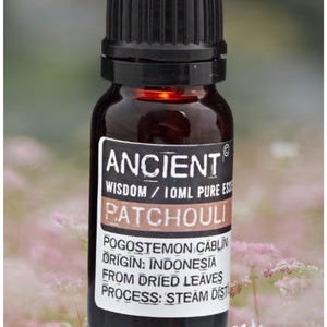 Op de afbeelding: Een bruine glazen fles patchouli etherische olie met een zwarte dop. Het etiket luidt "ANCIENT WISDOM / 10ML PURE ESSENTIAL OIL PATCHOULI". De fles staat tegen een vage bloemenachtergrond.