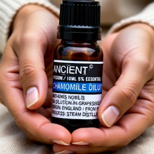 Peut inclure: Petite bouteille en verre ambré d'huile essentielle de camomille, tenue dans des mains jointes. L'étiquette indique "ANCIENT" et "CHAMOMILE DILUT". Le flacon a un bouchon noir. Le texte "Gentle Care for Sensitive Souls" est en bas.