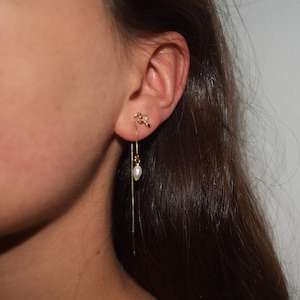 Könnte beinhalten: Ein goldener Kettenohrring mit einem kleinen Perlenanhänger. Der Ohrring wird am linken Ohr einer Person mit langen braunen Haaren getragen.