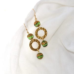 Könnte beinhalten: Goldene Ohrringe mit grünen Glasperlen und strukturierten runden Anhängern.