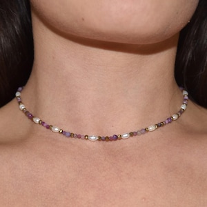 Könnte beinhalten: Eine zarte Choker-Halskette mit einer Kette aus kleinen, glänzenden Perlen, die mit lila und goldfarbenen Perlen durchsetzt sind. Die Halskette ist so konzipiert, dass sie eng am Hals anliegt und ein subtiles und elegantes Accessoire bietet.
