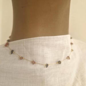 Könnte beinhalten: Zarte goldene Halskette mit kleinen, unregelmäßig geformten Edelsteinen in Rosa-, Braun- und Grautönen. Die Halskette ist an einer feinen Goldkette aufgereiht und vor einem weißen Stoffhintergrund zu sehen.