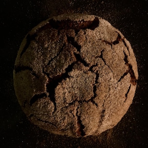 May include: A close-up of a round, dark brown loaf of bread with a cracked crust. The bread is covered in a fine layer of flour.
