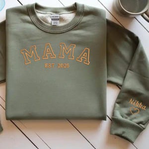 Könnte beinhalten: Militärgrünes Sweatshirt mit goldfarbenem "MAMA" und "EST 2026" gestickt. Der Ärmel zeigt den Namen "Nikka" und ein Herz. Eine Tasse und Blätter sind ebenfalls zu sehen. Ein bequemes Kleidungsstück.