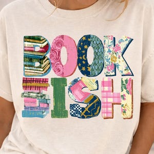 May include: Cream-colored t-shirt with the word "BOOKISH" in colorful, patterned letters. The letters are decorated with images of books, roses, stars, and floral designs. A stylish and unique graphic tee for book lovers.
