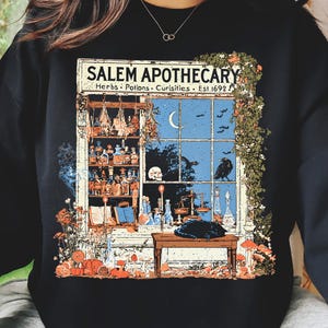 以下が含まれることがあります： SALEM APOTHECARY Herbs, Potions, Curiosities, Est. 1692と書かれた店の窓のグラフィックプリントが施された黒いスウェットシャツ。イラストには、ボトルの棚、頭蓋骨、カラス、三日月、木製のテーブルの上に座る黒猫が描かれています。
