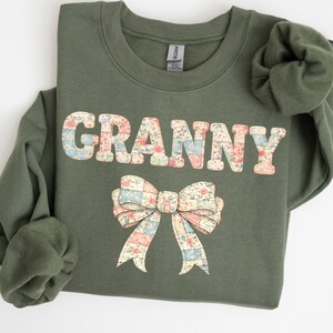 May include: An olive green sweatshirt with the word "GRANNY" in a floral patchwork design. A matching patchwork bow is below the word. The sweatshirt is folded, with the sleeves visible.