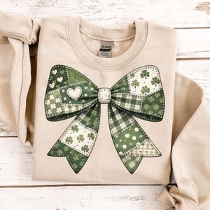 Può includere: Felpa beige con un grande design a fiocco patchwork. L'arco è principalmente verde con vari motivi, tra cui trifogli, cuori e quadri. Un cuore bianco è al centro. La felpa è appoggiata su una superficie di legno chiaro.