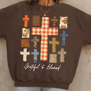 May include: A dark brown crewneck sweatshirt featuring a large central orange and white gingham cross surrounded by smaller crosses in various patterns including leopard print, floral, plaid, and denim. The text Grateful & Blessed is printed below.