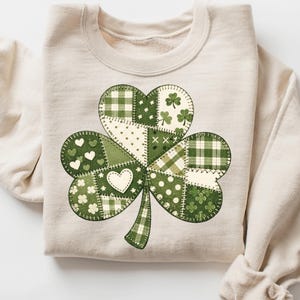 以下が含まれることがあります： ベージュのスウェットシャツ。大きなクローバーのデザインが特徴です。クローバーは、水玉模様、ハート、チェック柄など、さまざまな緑色のパッチワークで構成されています。デザインはスウェットシャツの前面に縫い付けられています。