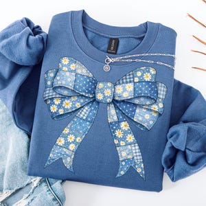May include: A blue sweatshirt with a large bow design. The bow is made of patchwork fabric in shades of blue, white, and yellow, with a daisy pattern. A silver necklace with a small pendant is also visible.