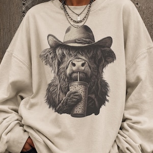 Op de afbeelding: Cr&egrave;mekleurige sweatshirt met een zwart-wit illustratie van een Schotse hooglander met een cowboyhoed en drinkend uit een beker met patroon. De vacht van de koe is gedetailleerd en de hoed heeft een klassieke vorm. De sweatshirt heeft een ronde hals en lange mouwen.