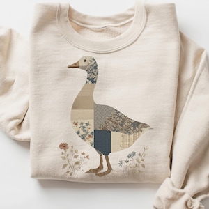 May include: A cream-colored sweatshirt featuring a patchwork goose design. The goose is composed of various patterned fabrics in shades of beige, blue, and gray. Floral accents and a brown beak complete the design. The sweatshirt has a crew neck and long sleeves.