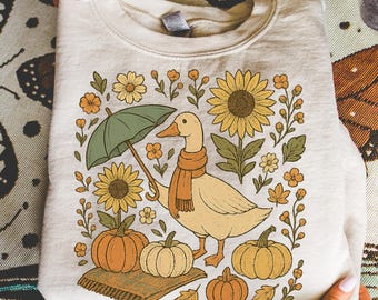May include: Cream-colored sweatshirt featuring a whimsical illustration of a duck wearing a scarf and holding an umbrella. The design includes sunflowers, pumpkins, and fall foliage. The sweatshirt is folded and held by hands.
